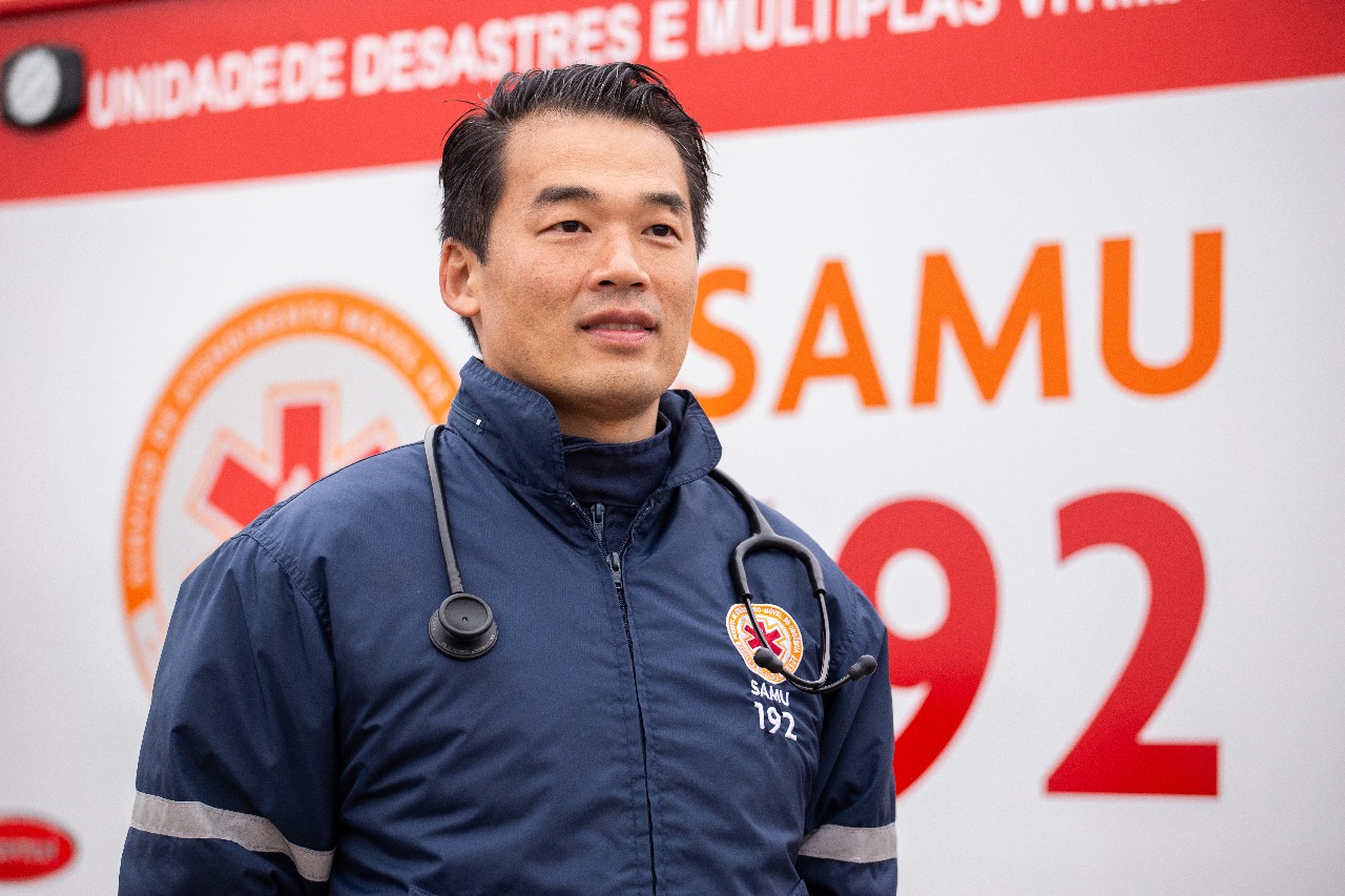 A foto mostra Francis, um homem japonês, de cabelos pretos, usando o uniforme do Samu. Ele está parado em frente e uma ambulância do serviço. 